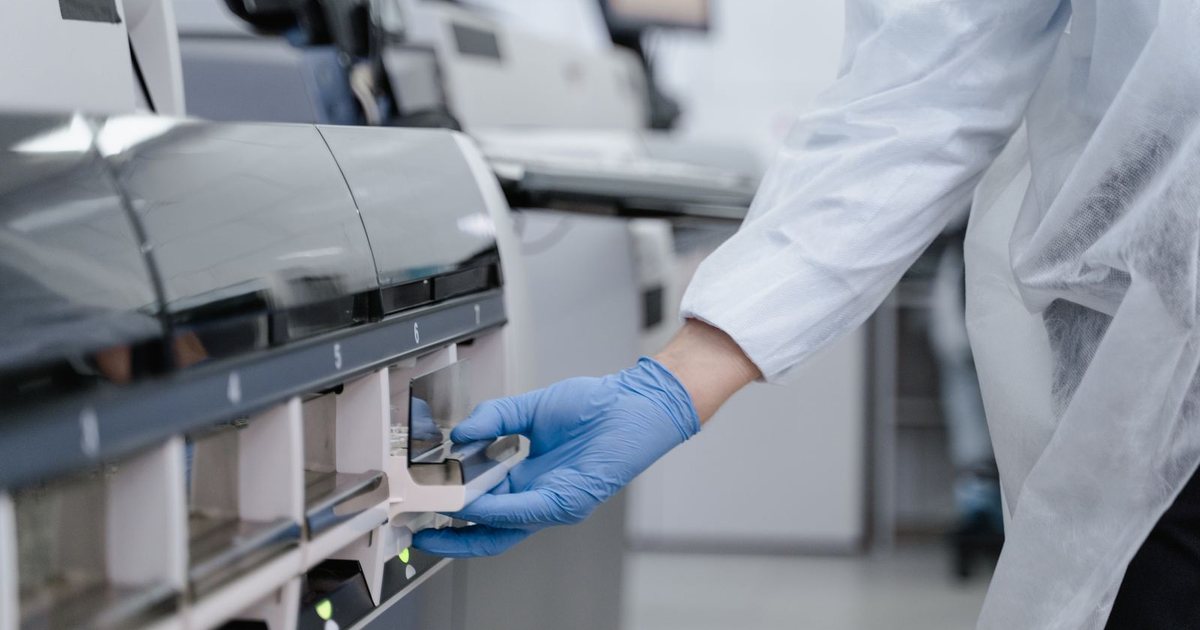 Mano enguantada manipulando un analizador de laboratorio en un entorno de sala blanca