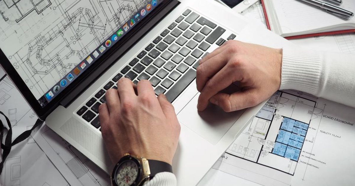 Manos escribiendo en un portátil junto a planos técnicos sobre un escritorio