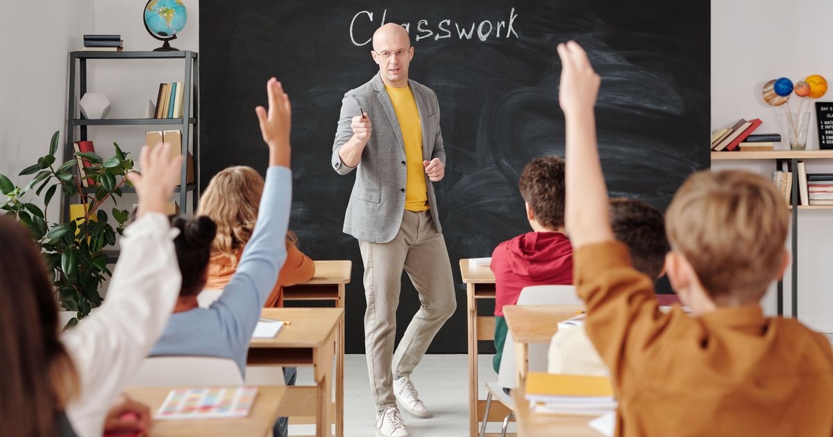 Profesor impartiendo una clase con la pizarra al fondo y alumnos levantando la mano