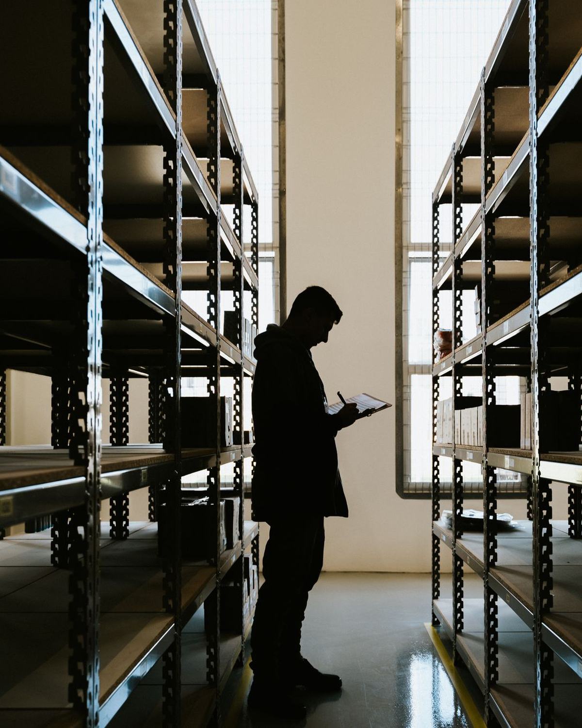 Silueta de una persona revisando inventario con una tableta entre estanterías metálicas vacías