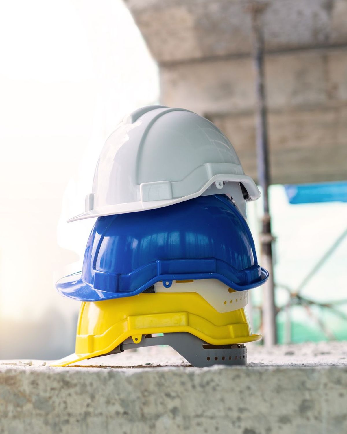 Tres cascos de obra apilados, blanco, azul y amarillo, sobre un bloque de hormigón