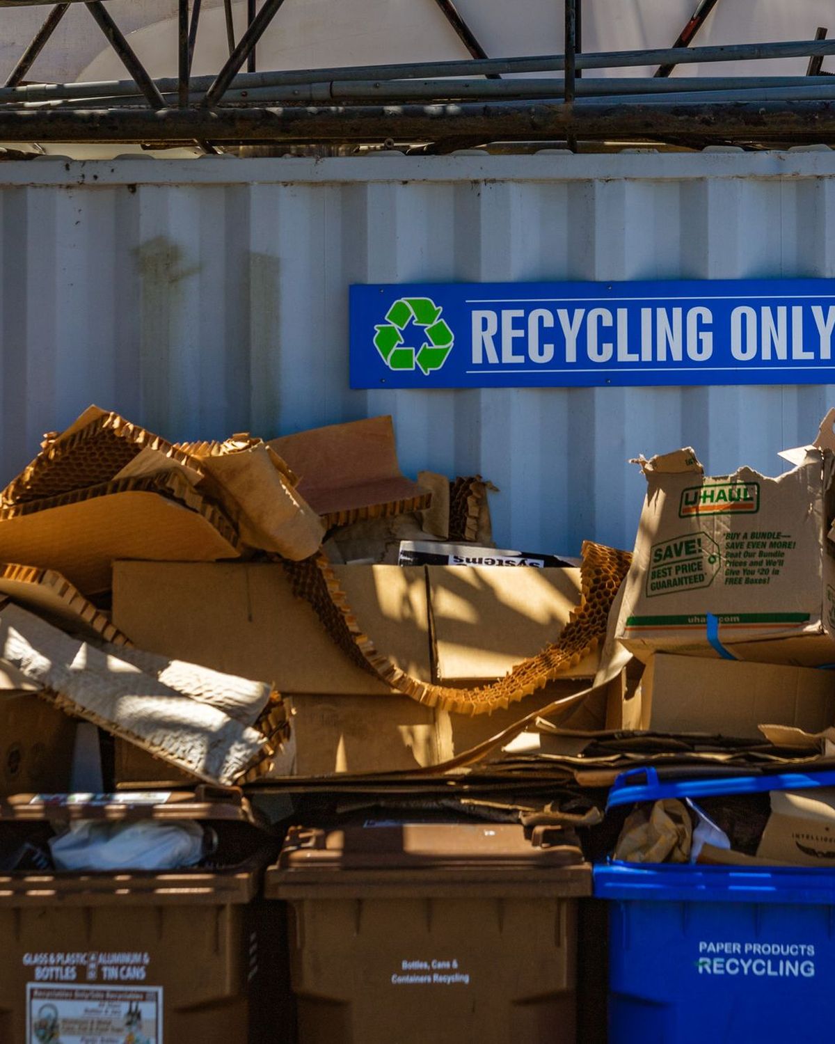 Contenedores de reciclaje con cartón y papel junto a un cartel de reciclaje
